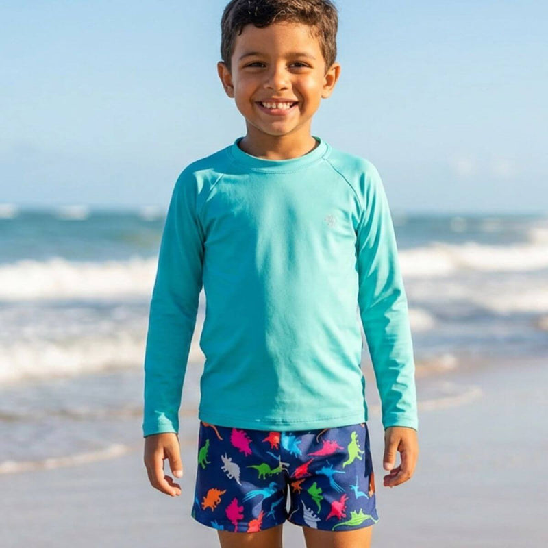 Menino sorrindo em pé na praia, vestindo uma camisa UV de manga longa azul claro e sunga estampada com dinossauros coloridos