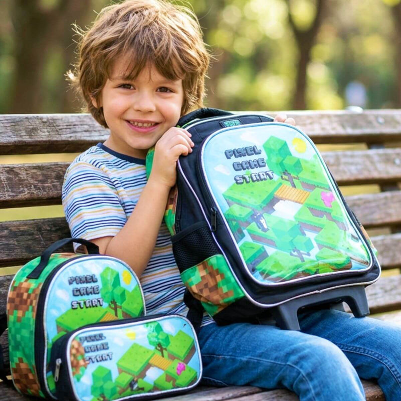 Menino sorrindo sentado em banco de parque segurando uma mochila de rodinhas preta com estampa verde de jogo pixelado