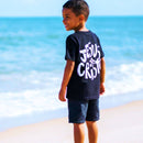 Criança sorrindo na praia usando camiseta branca infantil com pequeno logo de Jesus Cristo no peito.